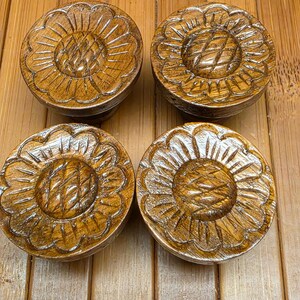 May include: A set of four circular wooden knobs, each featuring a carved sunflower design with a textured center and radiating petals. The wood has a polished, medium-brown finish with visible grain patterns throughout the surface.