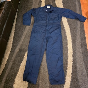 May include: A solid navy blue long-sleeve coverall featuring a collared neckline, a front button-up closure, and two chest pockets. The garment is constructed with a straight-leg design and long sleeves, providing full-body coverage in a uniform color.