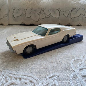 May include: A cream-colored toy car model featuring a sleek body, a hood scoop, and dark gray wheels. The vehicle is mounted on a rectangular dark blue plastic base, showcasing a classic coupe design with a light blue tinted windshield and windows.