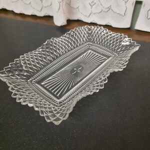 May include: A rectangular clear glass serving tray featuring a raised diamond pattern along the scalloped edges and a central sunburst design radiating from a small oval center. The glass has a textured finish and a reflective surface.