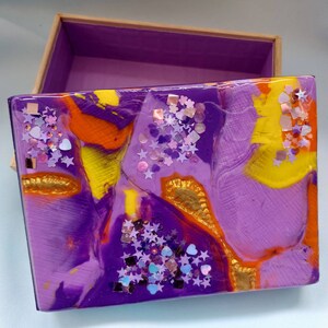 May include: A purple box with a lid decorated with yellow, orange, and gold swirls and glitter. The box is decorated with purple, pink, and blue glitter stars and hearts.