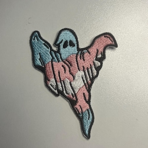 May include: Embroidered patch featuring a ghost design in the colors of the transgender pride flag: light blue, pink, and white. The ghost has a simple, friendly face and flowing form. This patch is suitable for jackets, bags, or other fabric items.
