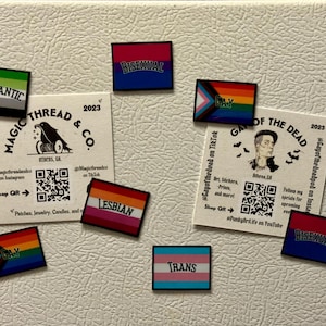 Może przedstawiać: Kolekcja magnesów i kart z flagami LGBTQ+, w tym flagi "Biseksualny", "Lesbijski", "Transpłciowy" i "Gay". Zawiera również karty z kodami QR i tekstem dla "Magic Thread & Co." i "Gay of the Dead".