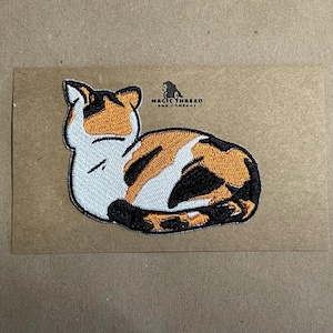 May include: A calico cat curled up in a sleeping position. The cat is white, orange, and black. The cat is embroidered on a brown fabric patch with the text "Magic Thread and Company" in black.