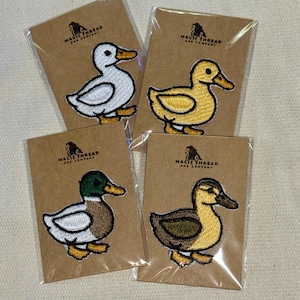 Op de afbeelding: Vier geborduurde eendenpatches in wit, geel en bruin, elk op een bruine kaart met de tekst "MAGIC THREAD AND COMPANY". De eenden hebben oranje snavels en gele poten. De patches zitten in doorzichtig plastic.