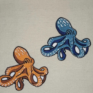 Pode incluir: Dois patches bordados de polvo, um azul e outro laranja, com detalhes brancos. Os patches estão sobre um fundo de tecido bege.