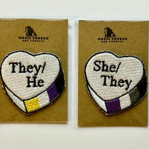 May include: Two heart-shaped embroidered patches, one with "They/He" and the other "She/They". Each has a white border and a non-binary flag design. They are displayed on brown cardstock, with the "Magic Thread and Company" logo.