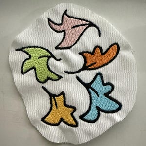 May include: A white fabric patch with five embroidered leaves in different colors: pink, green, orange, yellow, and blue.