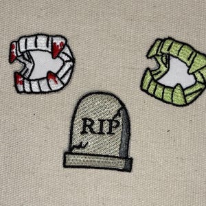 May include: Three embroidered patches on a beige background. One patch depicts a set of vampire fangs with red accents. Another shows green monster teeth. The third patch is a tombstone with the letters "RIP".