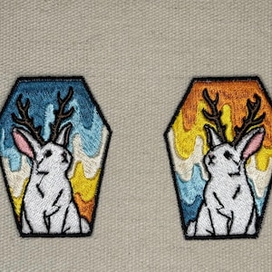 May include: Two embroidered patches featuring a white rabbit with antlers inside a hexagonal frame. One patch has a blue and yellow background, the other has an orange and yellow background. The rabbit has pink ears and a black outline.