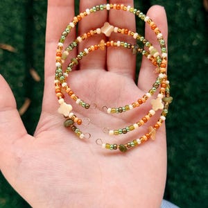 Könnte beinhalten: Ein Set aus vier Armbändern mit Perlen in Grün-, Orange- und Goldtönen. Jedes Armband hat kleine Perlen und sternförmige Akzente. Die Armbänder werden auf einer Hand präsentiert.