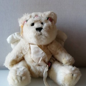 May include: A white plush teddy bear with wings and a satin ribbon. The bear has pink flowers on its head and a pink flower on its chest.