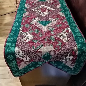 May include: A quilted table runner with a green border and a diamond pattern in shades of red, white, and brown. The runner features a floral print with green leaves and red flowers.