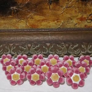 Peut inclure: Dix boutons de tiroir en céramique en forme de fleur avec un motif rose et jaune. Les boutons ont un centre blanc avec un tourbillon jaune et un bord extérieur rose.