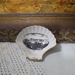 May include: A white porcelain shell-shaped trinket dish with a gold-trimmed edge. The center features a black and white illustration of the Percé Rock in Quebec, Canada, with the text "PERCE, P.Q. CANADA" printed clearly at the bottom.