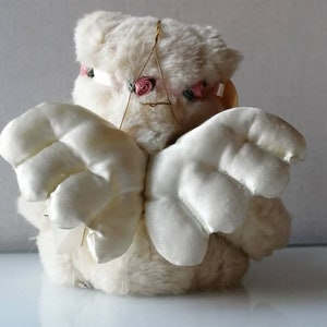May include: White plush angel with wings made of white satin. The angel has a pink flower headband.