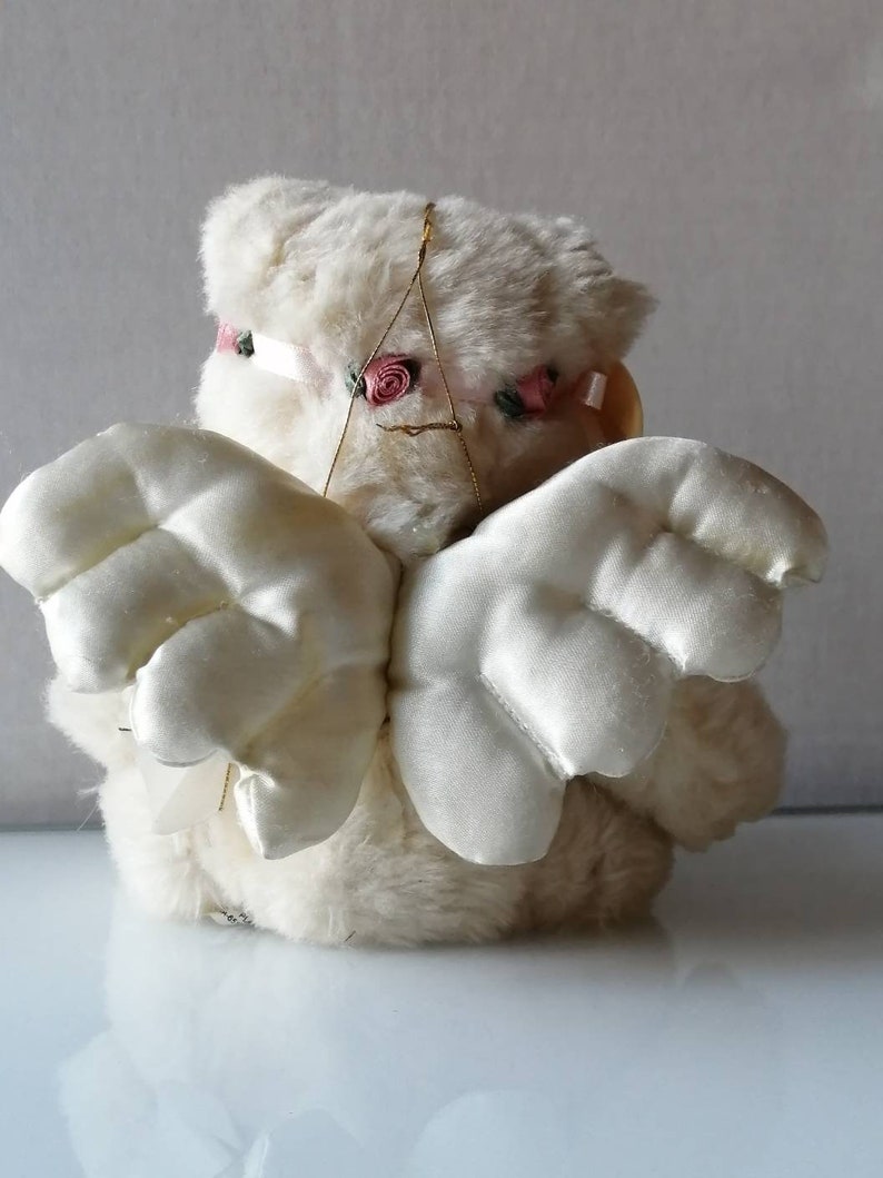May include: White plush angel with wings made of white satin. The angel has a pink flower headband.