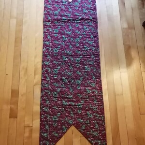 May include: A long, burgundy table runner with a green floral pattern. The runner is quilted and has a pointed shape at both ends.