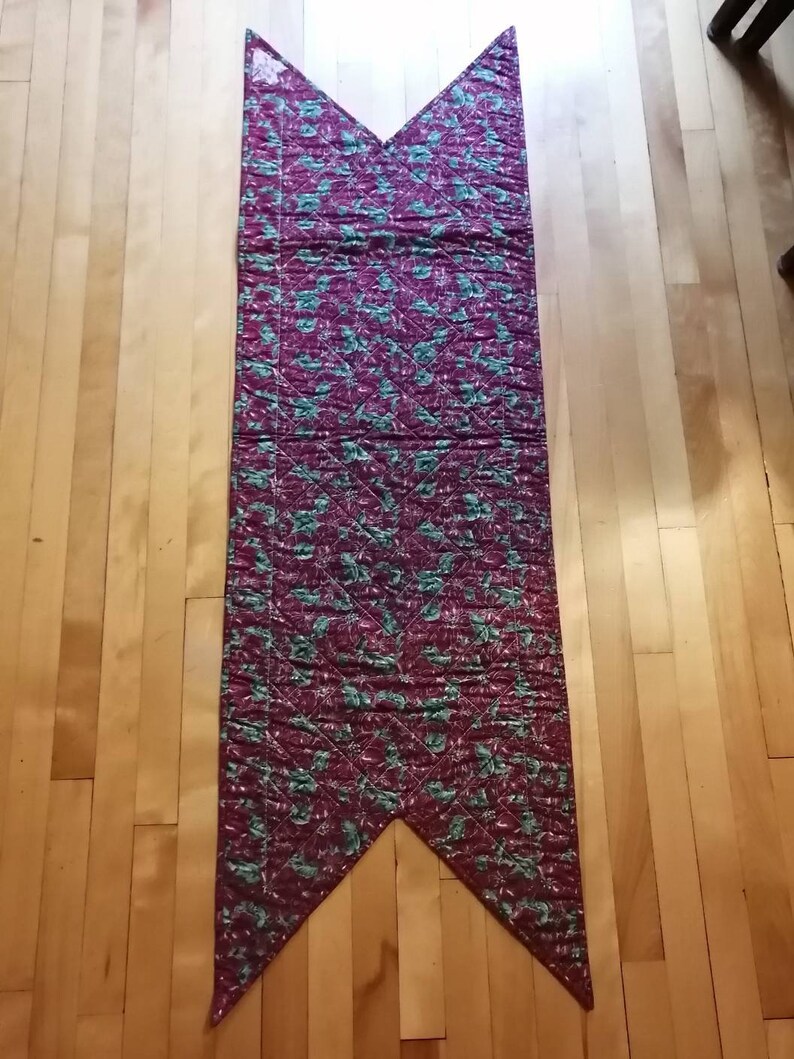 May include: A long, burgundy table runner with a green floral pattern. The runner is quilted and has a pointed shape at both ends.