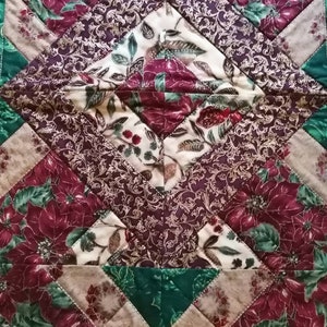 May include: A quilted table runner with a geometric pattern featuring red poinsettia flowers, green leaves, and gold accents on a brown and cream background.
