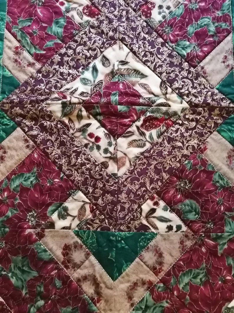 May include: A quilted table runner with a geometric pattern featuring red poinsettia flowers, green leaves, and gold accents on a brown and cream background.