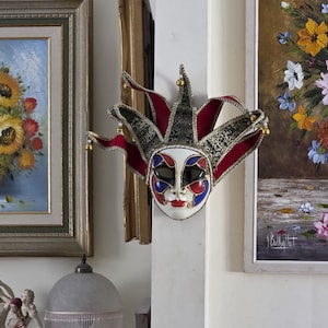 May include: A decorative Venetian mask in a jester style. The mask has a white face with red lips, blue and red eye details, and a red and black patterned headpiece with gold bells. It is hanging on a white wall.