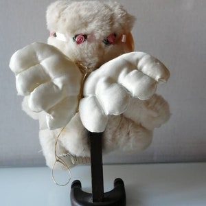 May include: A white plush angel with pink rose accents on its head and wings. The angel is standing on a wooden base.