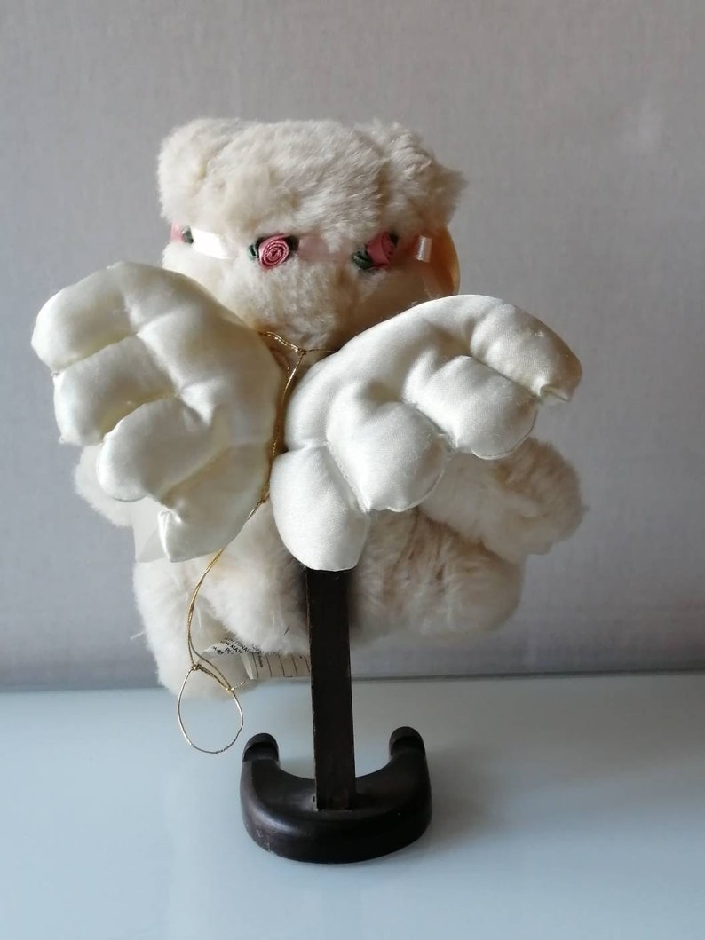 May include: A white plush angel with pink rose accents on its head and wings. The angel is standing on a wooden base.