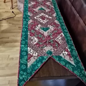 May include: A green and red quilted table runner with a diamond pattern. The runner has a pointed end and a green border with a floral design.