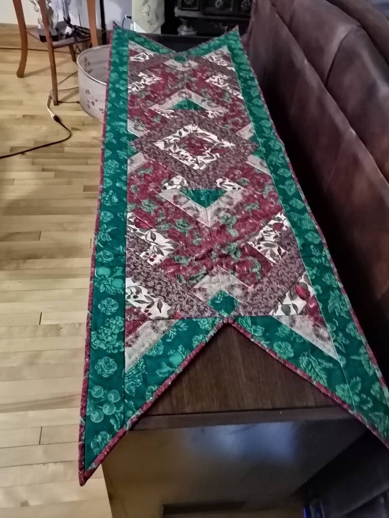 May include: A green and red quilted table runner with a diamond pattern. The runner has a pointed end and a green border with a floral design.