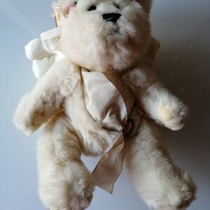 May include: A white plush teddy bear wearing a white satin ribbon and a white angel wing. The bear has pink accents on its ears and a black nose.