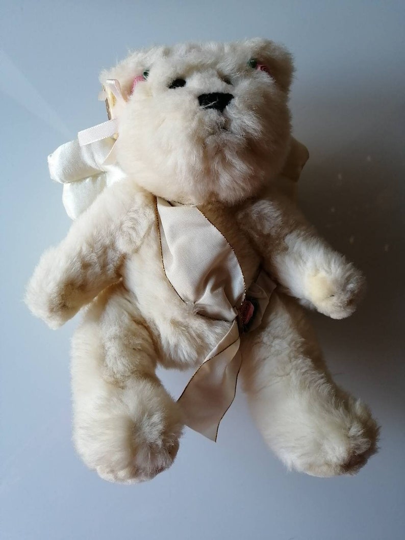 May include: A white plush teddy bear wearing a white satin ribbon and a white angel wing. The bear has pink accents on its ears and a black nose.