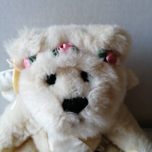 May include: A white plush teddy bear wearing a white satin dress and a flower crown with pink and green flowers. The bear has black eyes and a black nose.