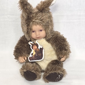 May include: A plush doll wearing a brown furry kangaroo costume with a hood and ears. The doll has a tag with a picture of a baby in the same costume. The doll is sitting on a white surface.