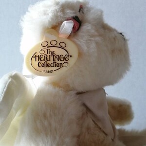 May include: White plush teddy bear with wings, wearing a pink bow and a white collar. The bear has a tag that reads "The Heritage Collection Ganz".