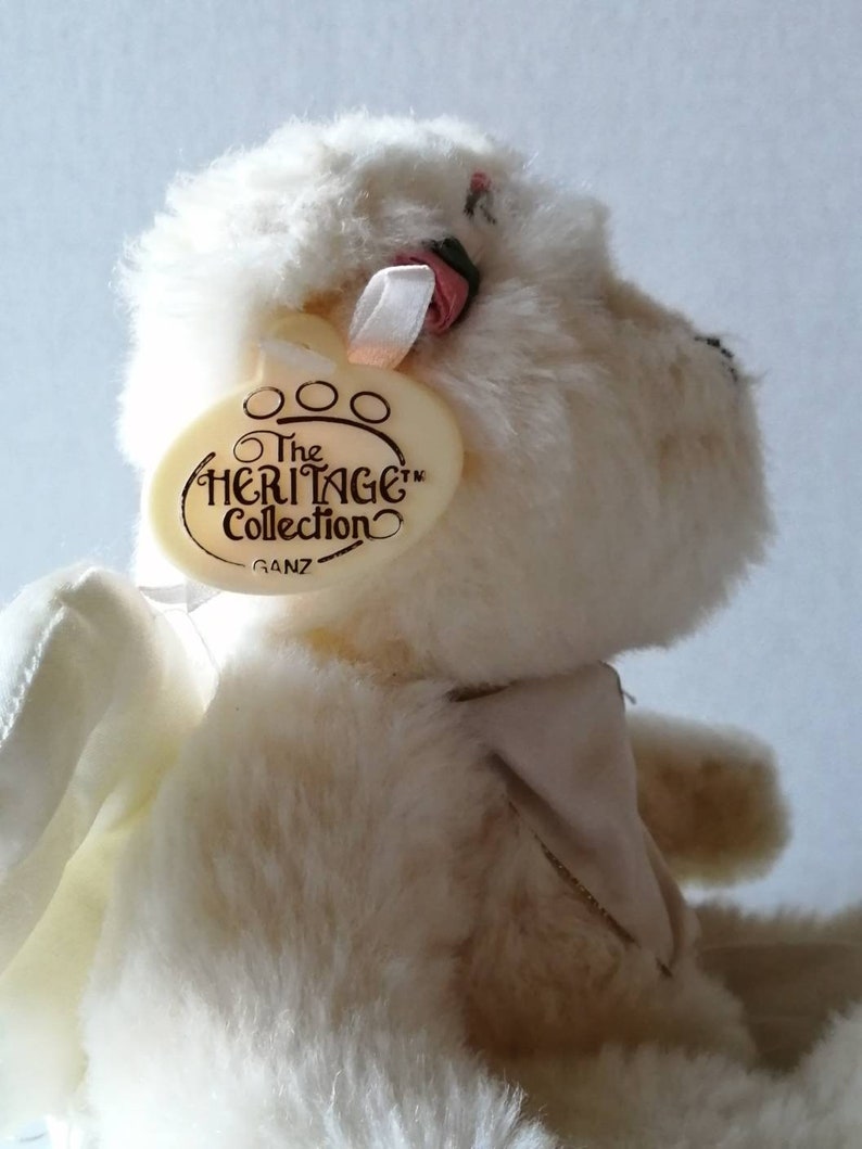 May include: White plush teddy bear with wings, wearing a pink bow and a white collar. The bear has a tag that reads "The Heritage Collection Ganz".