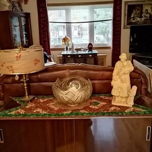 May include: A vintage wooden cabinet with two sliding doors, a green and brown patterned runner, a crystal bowl, a lamp with a floral patterned shade, and a white porcelain figurine of a woman holding a child.