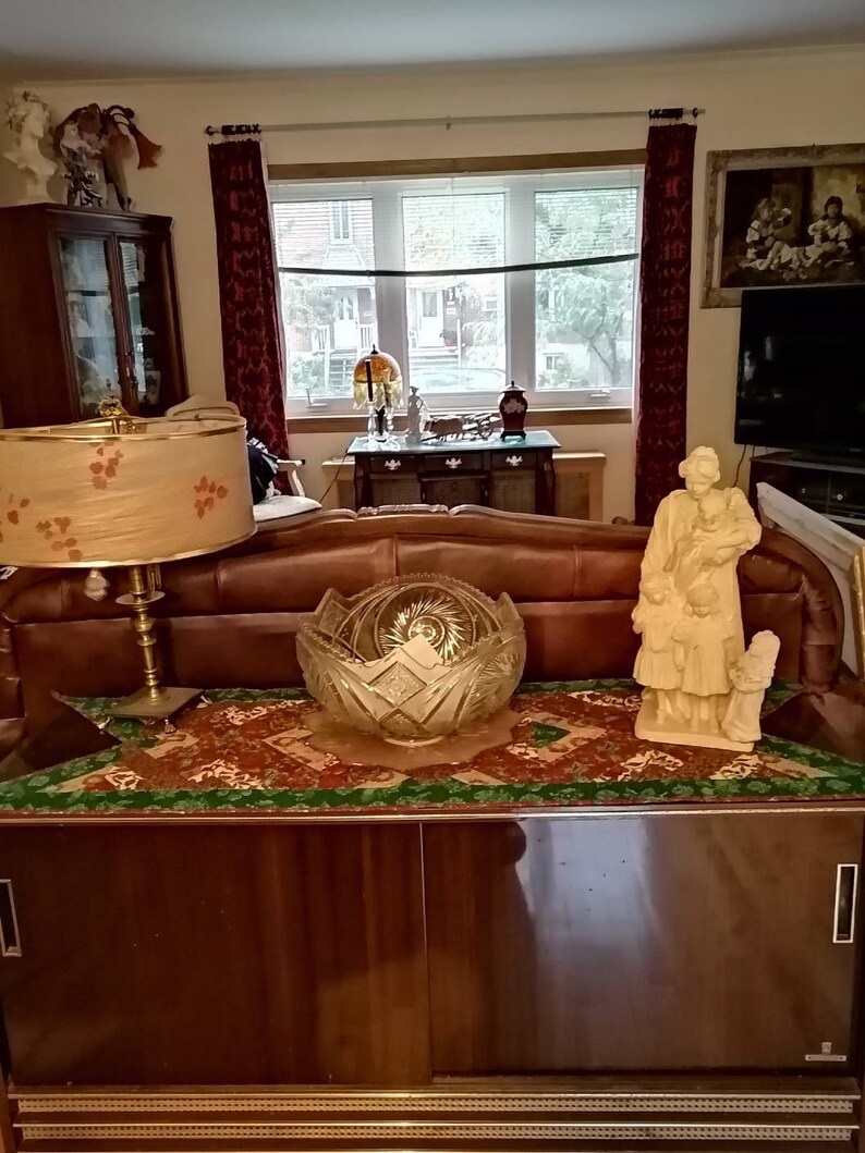May include: A vintage wooden cabinet with two sliding doors, a green and brown patterned runner, a crystal bowl, a lamp with a floral patterned shade, and a white porcelain figurine of a woman holding a child.