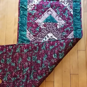 May include: A quilted table runner with a green border and a patchwork design featuring red, white, and green floral patterns.
