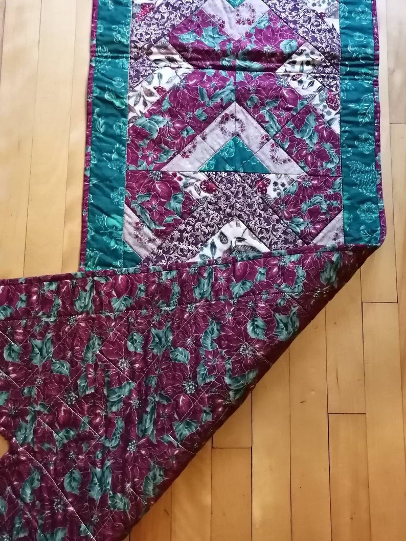May include: A quilted table runner with a green border and a patchwork design featuring red, white, and green floral patterns.