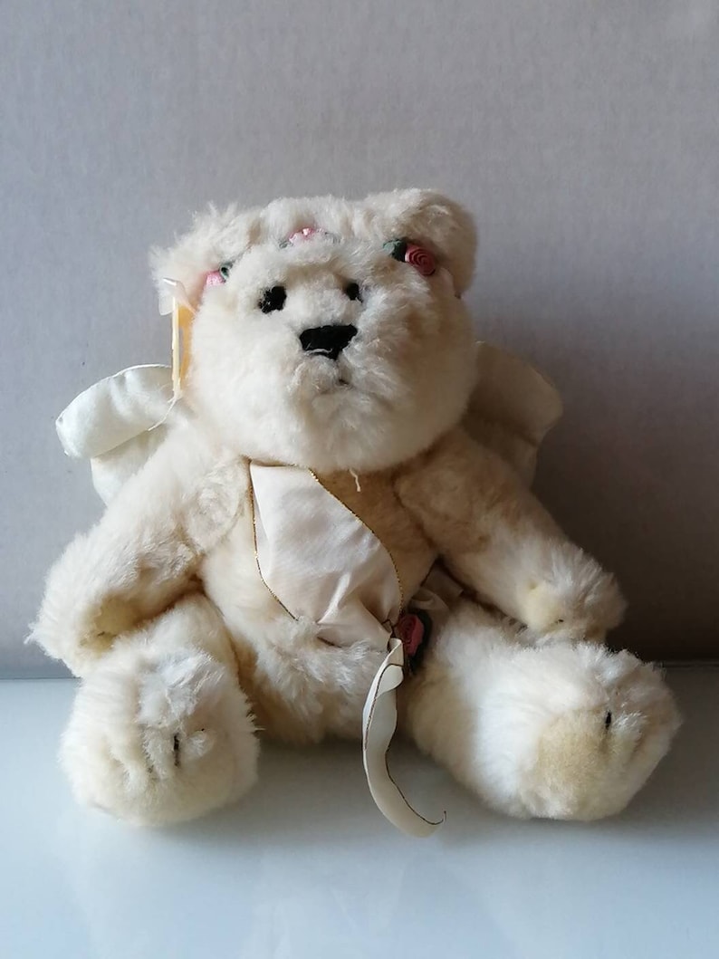 May include: A white plush teddy bear with wings and a satin ribbon. The bear has pink flowers on its head and a pink flower on its chest.