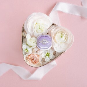 May include: A heart-shaped box filled with white and peach flowers, with a purple ring box containing a diamond ring in the center.