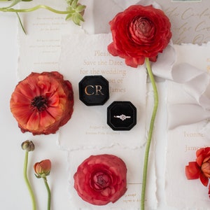 K&ouml;nnte beinhalten: Eine Hochzeitseinladungssuite mit roten Ranunkelblumen, einem Diamantring in einer schwarzen Samtbox und Save-the-Date-Karten. Die Karten haben goldenen Text auf strukturiertem wei&szlig;em Papier. Das Bild hat eine romantische, elegante Ausstrahlung.