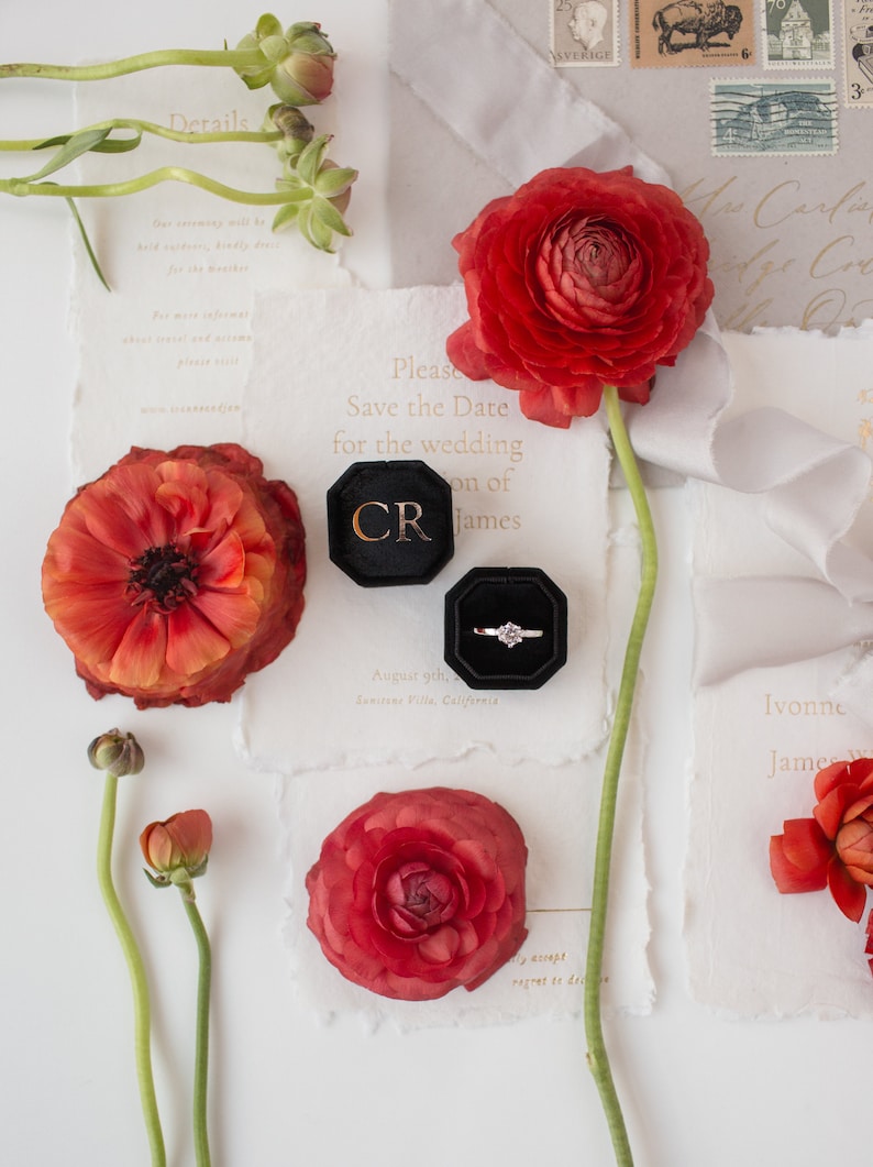 K&ouml;nnte beinhalten: Eine Hochzeitseinladungssuite mit roten Ranunkelblumen, einem Diamantring in einer schwarzen Samtbox und Save-the-Date-Karten. Die Karten haben goldenen Text auf strukturiertem wei&szlig;em Papier. Das Bild hat eine romantische, elegante Ausstrahlung.