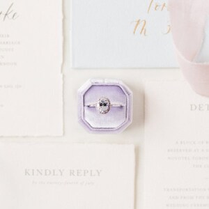 May include: A purple velvet ring box with a diamond engagement ring inside. The ring box is on a white surface with a white card that says "Kindly Reply" in grey text.