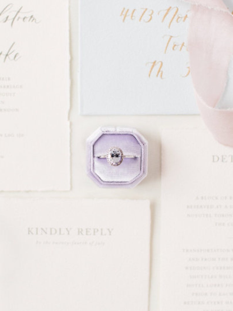 May include: A purple velvet ring box with a diamond engagement ring inside. The ring box is on a white surface with a white card that says "Kindly Reply" in grey text.