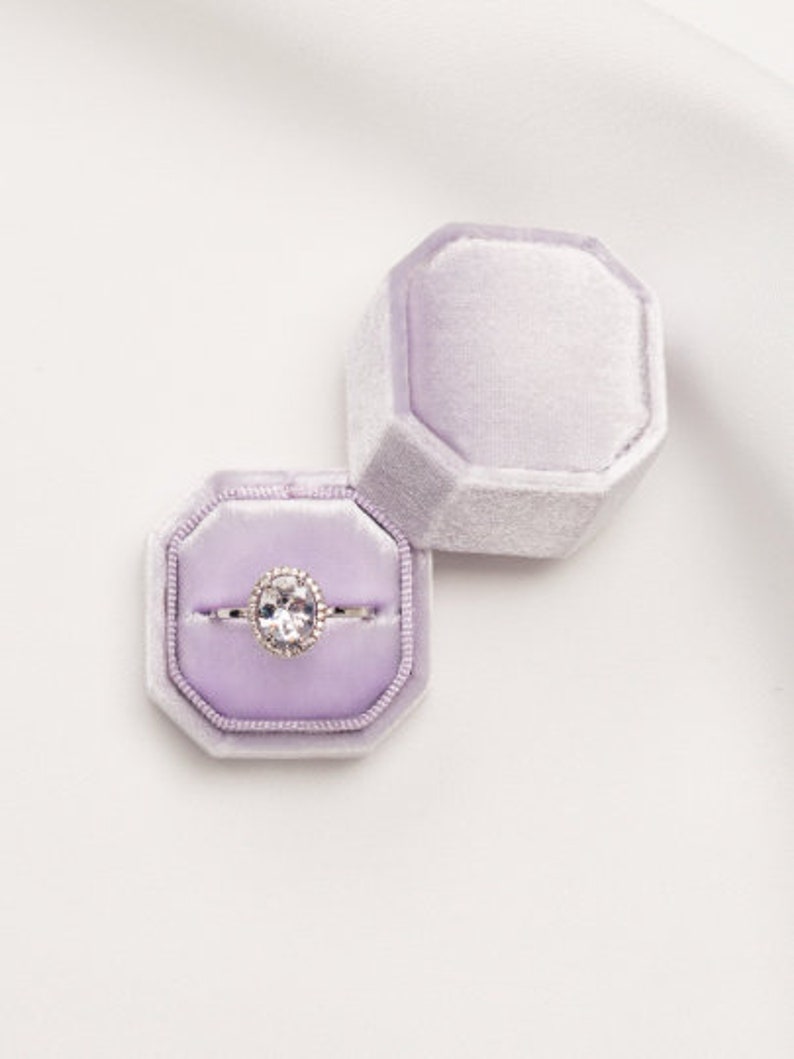 May include: A ring box with a lavender velvet interior. The box is open and a silver ring with a large oval-shaped diamond is inside.