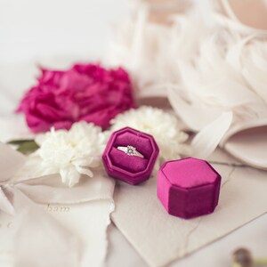 K&ouml;nnte beinhalten: Ein Diamant-Verlobungsring in einer magenta Samt-Ringbox, mit einer passenden leeren Box daneben. Wei&szlig;e Blumen, eine magenta Pfingstrose und wei&szlig;e High Heels mit Federakzenten im Hintergrund.