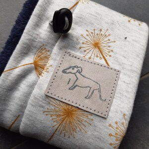 Könnte beinhalten: Weißer Stoff-Halswärmer mit schwarzem Knebelverschluss und einem kleinen braunen Patch mit einer weißen Strichzeichnung eines Hundes. Der Stoff hat ein sich wiederholendes Muster aus gelben Löwenzahnblüten.