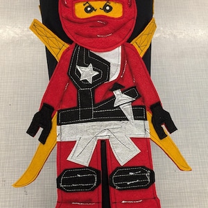 Könnte beinhalten: Eine rot-schwarze Ninja-Figur aus Filz mit gelben Akzenten. Die Figur hat eine rote Maske, schwarze Handschuhe und einen silbernen Stern auf der Brust. Zwei gelbe Katana-Schwerter sind hinter der Figur positioniert.